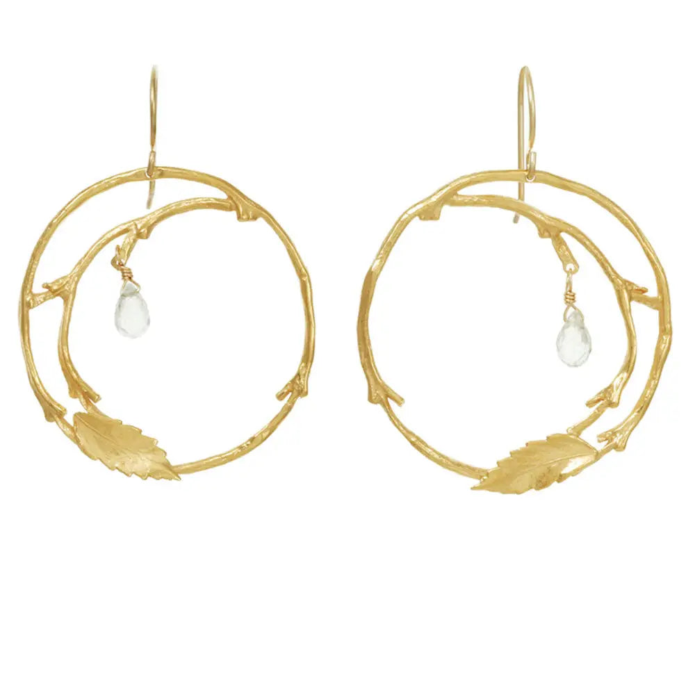 A pair of gold hoop earrings designed to look like organic tree branches with small leaf details and a clear teardrop crystal dangle, shown on a plain white background.