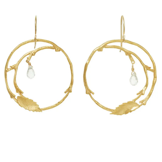 A pair of gold hoop earrings designed to look like organic tree branches with small leaf details and a clear teardrop crystal dangle, shown on a plain white background.